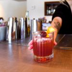 A bartender at the modern with tattoos on their forearm, gently pushes the Western Dynasty cocktail across the 3form countertop towards the photographer. The craft cocktail is a maroon color with a large clear ice cube and a twist of lemon served in a highball glass. The bar behind is stocked with typical bar tools like shakers, strainers, and other garnishes. On the wall behind the bartender you can see part of the liquor cabinet in the mid-century building in downtown boise idaho historic linen district.