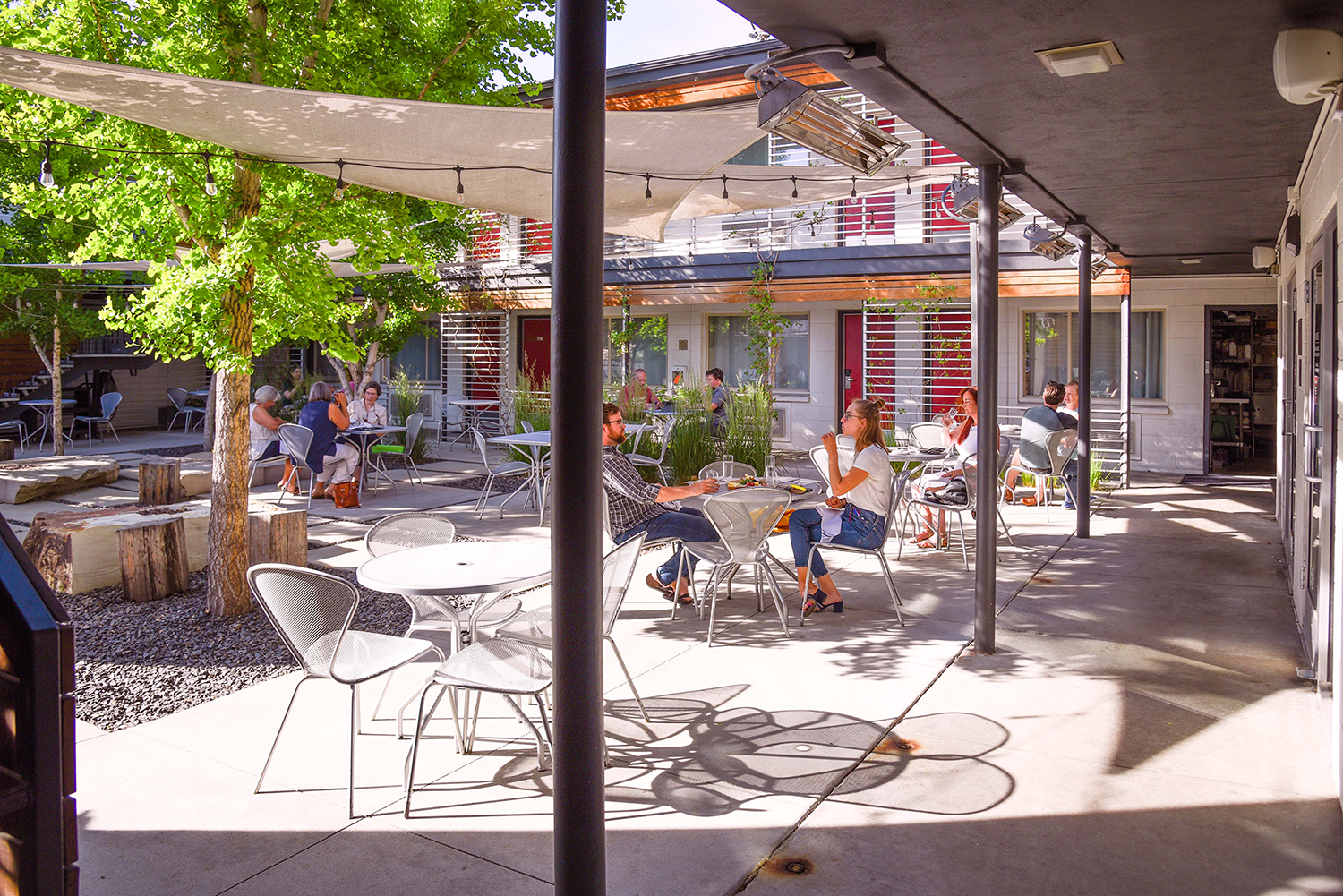 View of the Modern Hotel and Bar courtyard from the southeast corner of the patio. Several metal mesh tables with matching cherner-like chairs are occupied by patrons enjoying craft cocktails and appetizers. Reddish-orange hotel room doors are visible surrounding the courtyard. Ginkgo trees and sails provide shade.