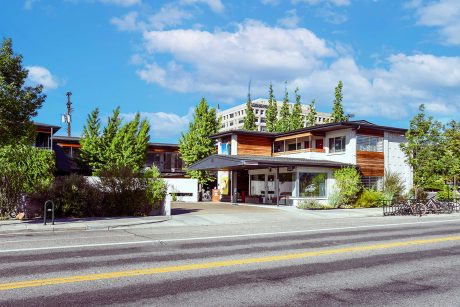 A view of the Modern Hotel and Bar in downtown Boise Idaho. The hotel is a renovated two-story travelodge. The photo was taken across Grove Street, looking at the lobby entrance of the motor hotel. The mid-century architecture is complimented by wood and metal accents, ginkgo trees in the courtyard, and hackberry vines. The landscape is xeriscape and includes native plants. The Idaho Power office building top stories are just visible behind the hotel. Boise's typical summer blue sky is on full display with whisperings of white clouds. 